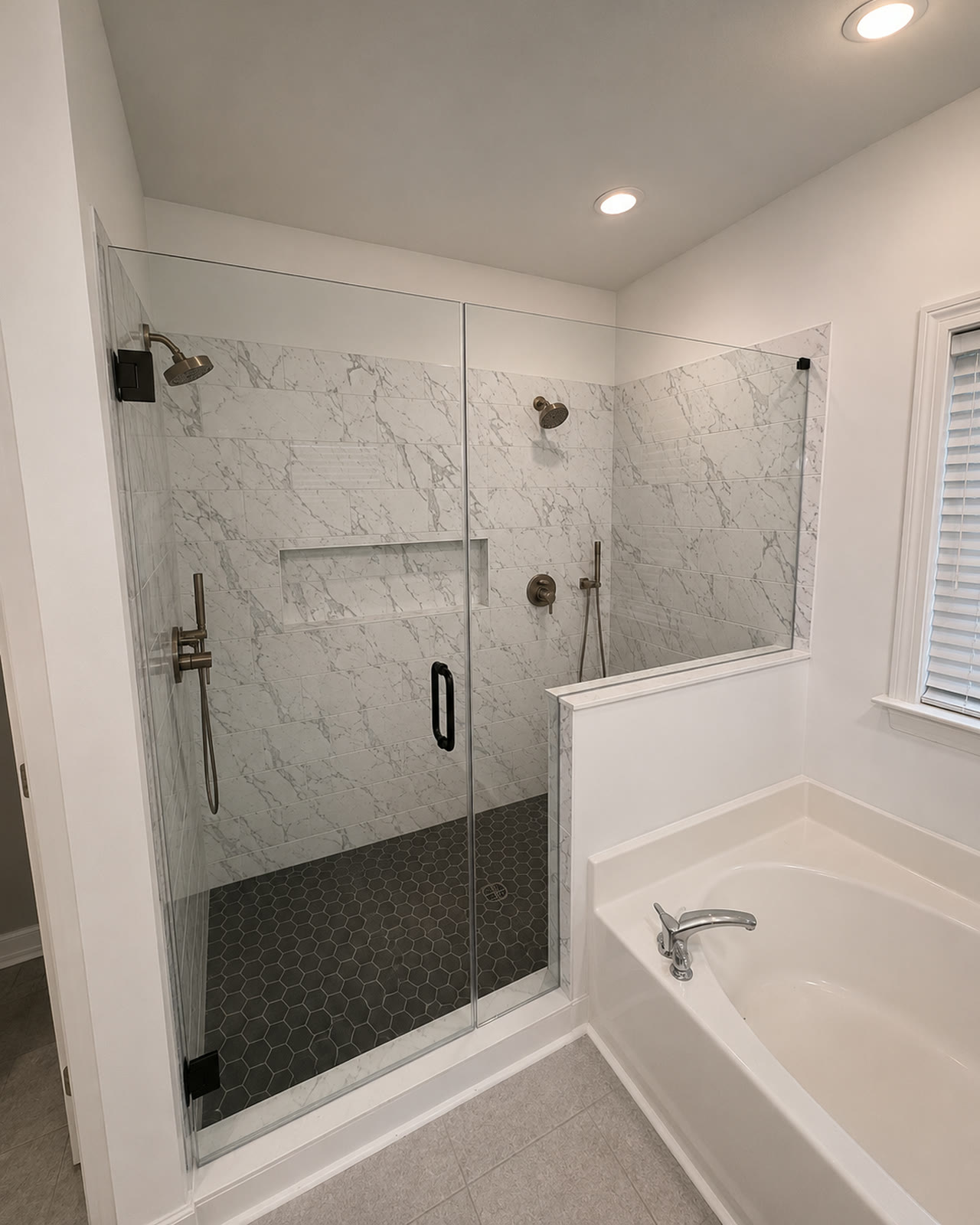 After: marble-look tile to the ceiling, a cut-in niche, black hex mosaic floor and frameless glass, shot from the same corner
